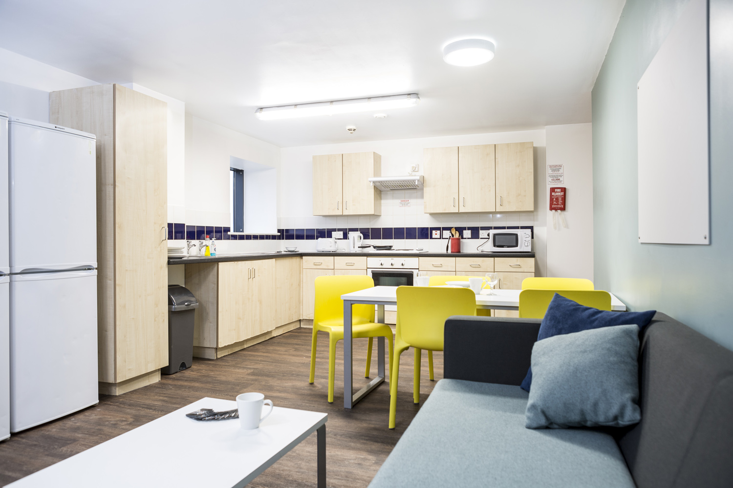 Living space at Arena Village, dark blue sofas in the foreground with brightly coloured chairs at the kitchen table