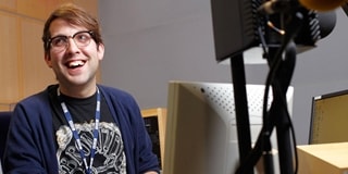 Man sat behind a computer and a speaker while smiling at the camera