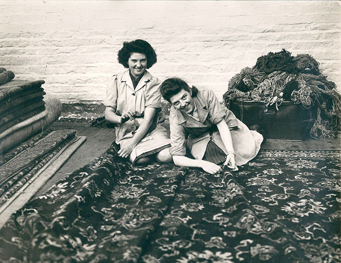 An old black and white photo showing two women working outside at Clifton Mill sewing a large carpet and smiling